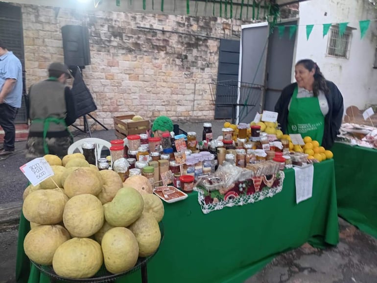 Mujer sonriente en delantal verde atiende un puesto de comida, con mesas verdes y productos variados en el mercado.