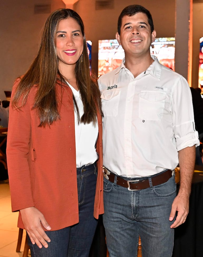 Mujer con blusa blanca y abrigo marrón junto a hombre con camisa blanca sonríe en ambiente decorado.