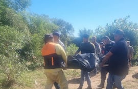 Seis individuos llevan un bulto cubierto por tela oscura, con chalecos de seguridad, en un área con vegetación.