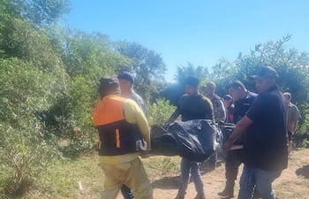 Seis individuos llevan un bulto cubierto por tela oscura, con chalecos de seguridad, en un área con vegetación.