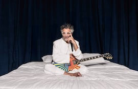 Artista sentado en una cama con sábanas blancas, vestido de blanco, guitarra colorida, expresión contemplativa en un ambiente oscuro.