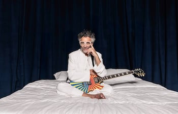 Artista sentado en una cama con sábanas blancas, vestido de blanco, guitarra colorida, expresión contemplativa en un ambiente oscuro.
