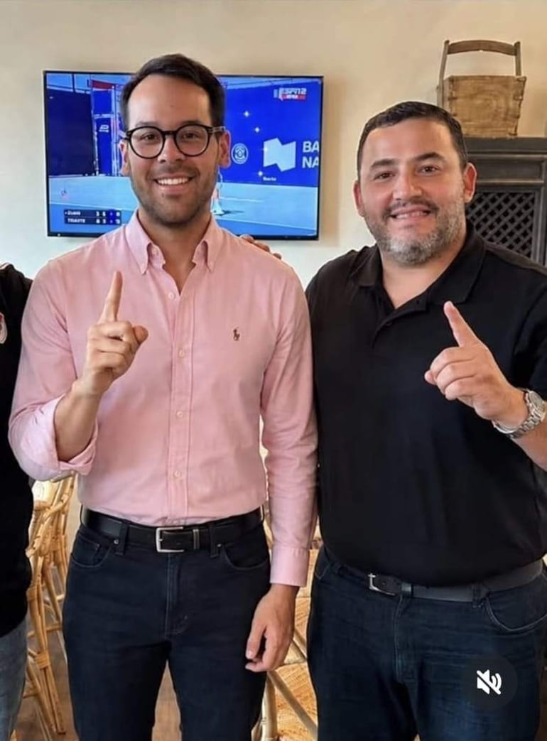 Axel Mongelós con camisa rosa y gafas, y Kike Chávez en camiseta negra, sonríen y hacen un gesto de celebración mientras un monitor los acompaña.