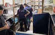 Dos mujeres con chalecos de Vigilancia Sanitaria revisan estantes mientras un hombre usa su teléfono móvil.
