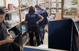 Dos mujeres con chalecos de Vigilancia Sanitaria revisan estantes mientras un hombre usa su teléfono móvil.