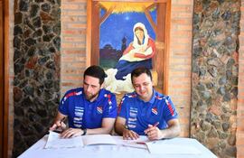 Gamarra y Ramírez en camisetas deportivas azules, firmando documentos en un ambiente acogedor con pintura religiosa al fondo.