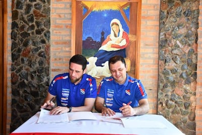 Gamarra y Ramírez en camisetas deportivas azules, firmando documentos en un ambiente acogedor con pintura religiosa al fondo.