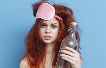 Una mujer pelirroja despeinada con una botella de agua en las manos y un antifaz para dormir mal puesto.