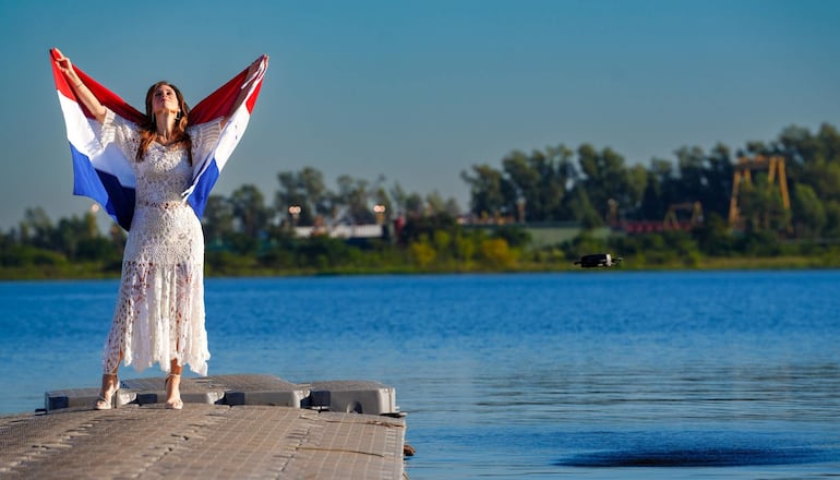 Mujer con vestido blanco de encaje y sandalias orgullosamente sostiene la bandera paraguaya en un muelle bajo un cielo despejado.