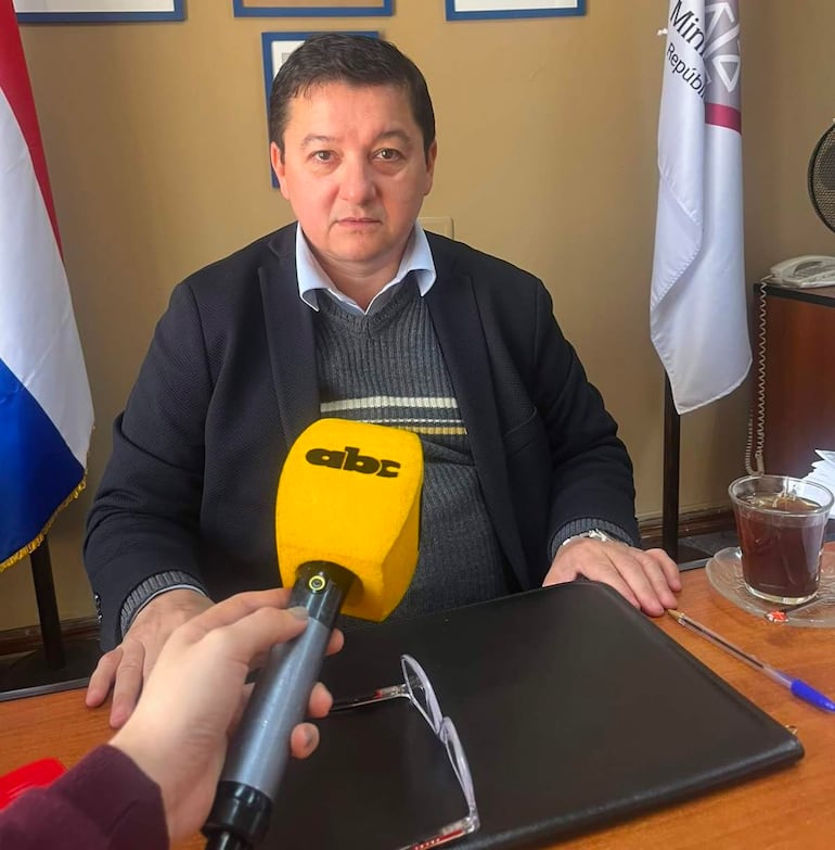 Julio Ortíz sentado en escritorio con suéter gris y saco negro, con micrófono amarillo sostenido por reportero.