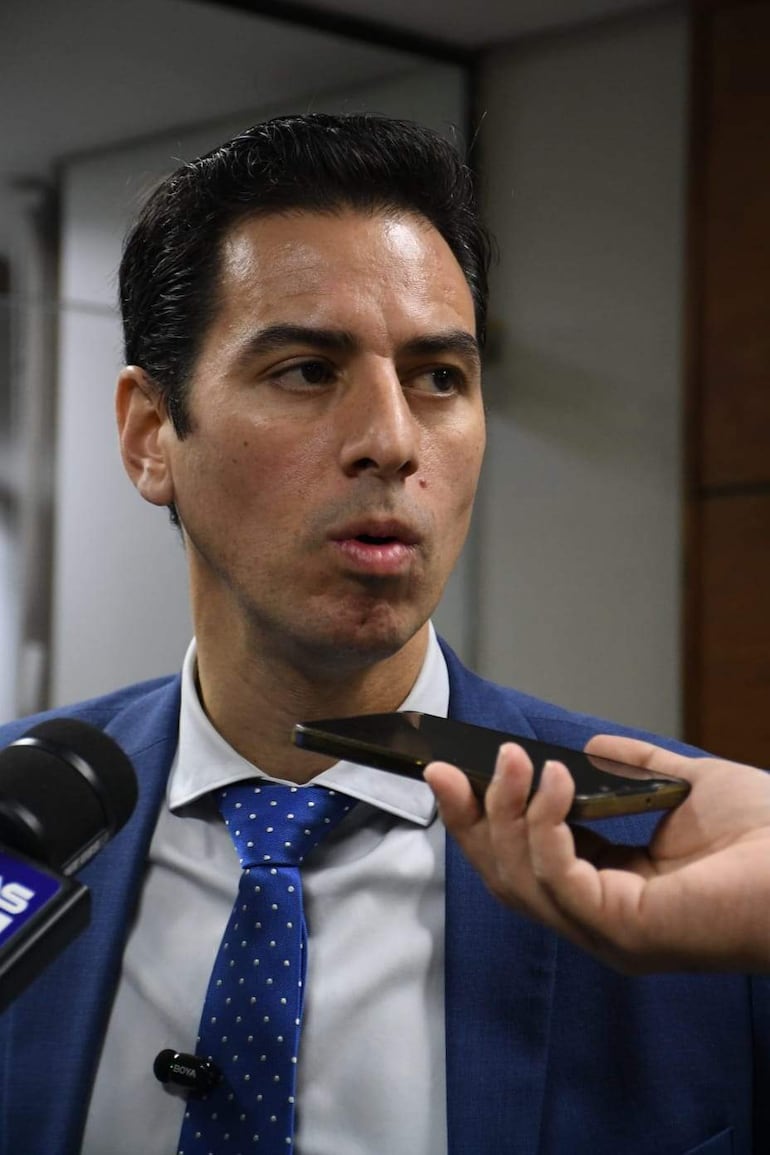 Alvaro Grau en primer plano con traje azul y corbata de puntos, sostiene un micrófono mientras un periodista graba con su teléfono.