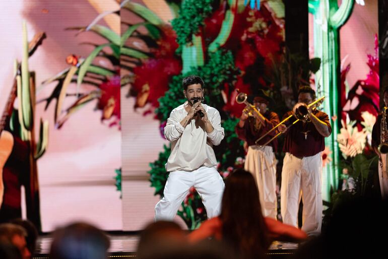 Cantante con camisa y pantalones blancos actúa en un escenario festivo, rodeado de músicos con camisas burdeos, público disfrutando.
