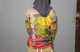 Persona con sudadera y paño oscuro en la cabeza, vista de espaldas, en interior con pared gris.