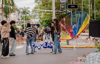 Mujer con abrigo marrón y jeans sosteniendo un micrófono, rodeada de jóvenes conversando en un ambiente festivo al aire libre.