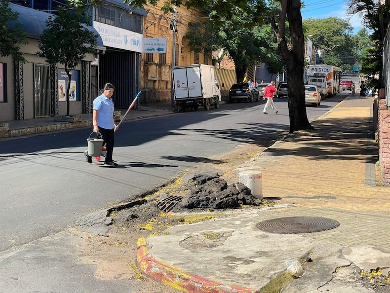 Hombre con camiseta clara y pantalones oscuros sostiene un balde y un palo de limpieza, con bache visible en el pavimento.