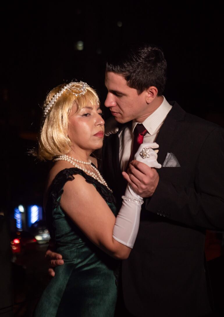 Mujer con vestido verde y guantes blancos junto a hombre en traje negro y corbata roja, en un ambiente nocturno con luces difusas.