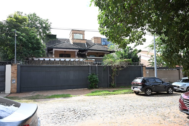 Casa de Dalia López con techo a dos aguas y vegetación, rodeada de una verja de alambre de púa y vehículos en la calle.