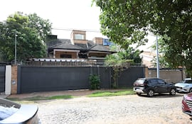 Casa de Dalia López con techo a dos aguas y vegetación, rodeada de una verja de alambre de púa y vehículos en la calle.