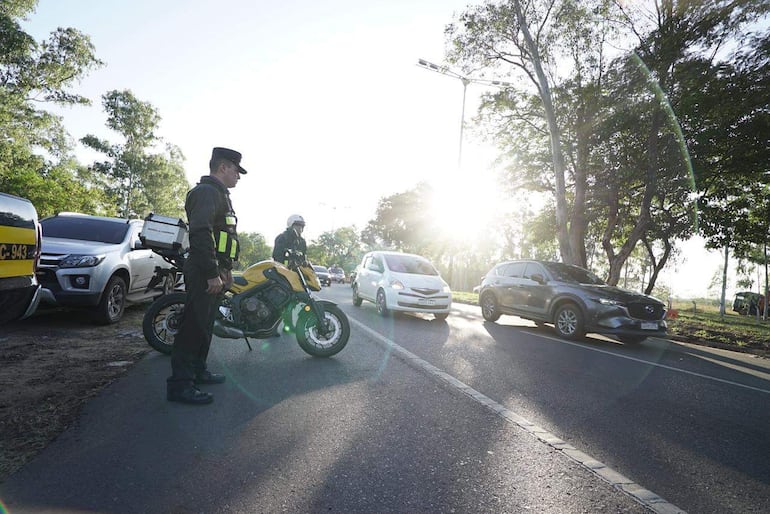 Dos policías en uniforme, uno montado en una motocicleta amarilla, vigilan el tráfico en la vía pública rodeada de árboles.