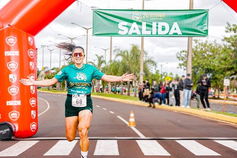 Mujer con camiseta verde y pantalones cortos cruza el arco de meta sonriente, rodeada de espectadores en un ambiente festivo.