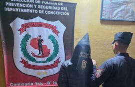 Hombre con saco negro, cabeza cubierta y postura sumisa junto a agente de policía en comisaría austera con paredes amarillas.