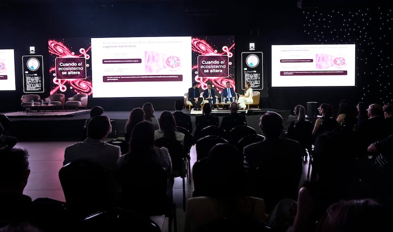 Panel de cuatro personas, incluyendo una mujer de blanco hablando. Pantallas con texto sobre salud íntima femenina al fondo.