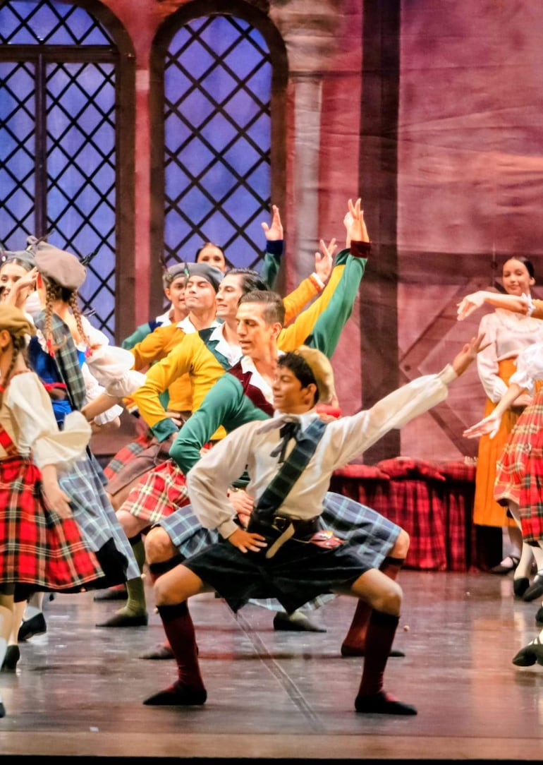 Bailarines en vestimenta escocesa, faldas de cuadros y camisas coloridas, bailan con alegría en escenario con fondo arquitectónico.