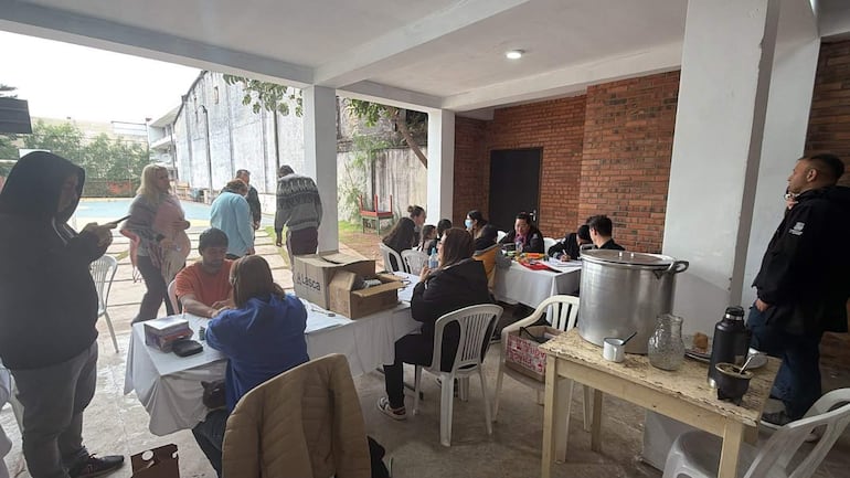Mujer en chaqueta azul conversando con hombre de camiseta naranja en mesa, rodeados de personas en un ambiente comunitario informal.