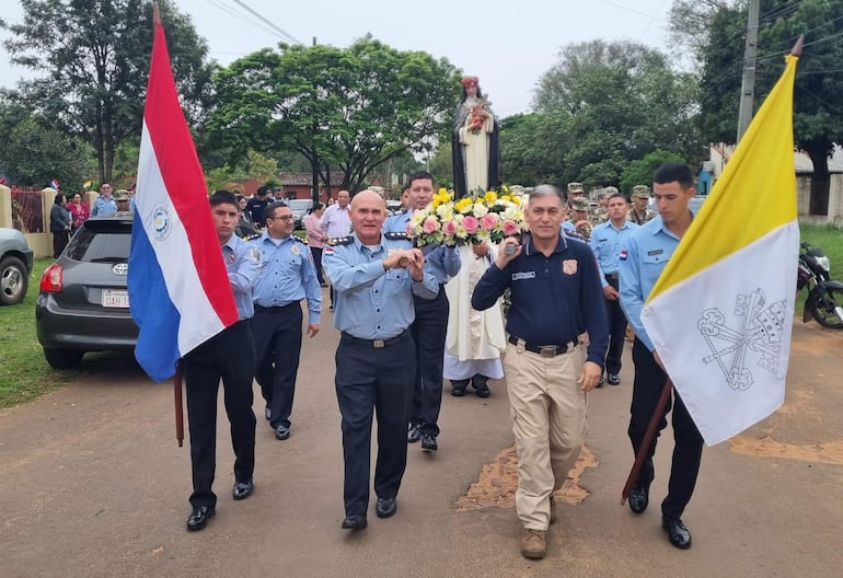 Hombres con banderas, marco floral y figura religiosa, seguidos por un sacerdote en vestimenta litúrgica durante la procesión.