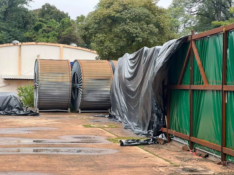 Bobinas metálicas cubiertas con lona negra en un área industrial con estructura curva al fondo, sin personas presentes.