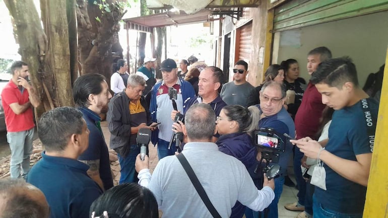 Hombre con micrófono al frente hablando, rodeado de personas con cámaras y teléfonos. Ambiente de protestas con árboles alrededor.