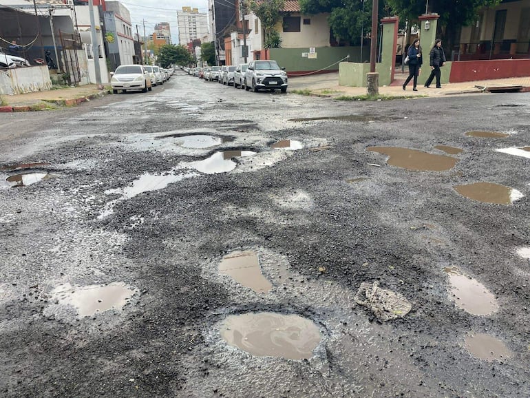 Dos personas con chaquetas caminan junto a vehículos blancos, mientras la calle muestra baches llenos de agua.
