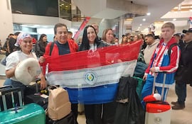 Un hombre y una mujer sostienen una bandera de Paraguay, rodeados de jóvenes felices en el aeropuerto con equipaje.
