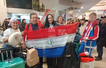Un hombre y una mujer sostienen una bandera de Paraguay, rodeados de jóvenes felices en el aeropuerto con equipaje.