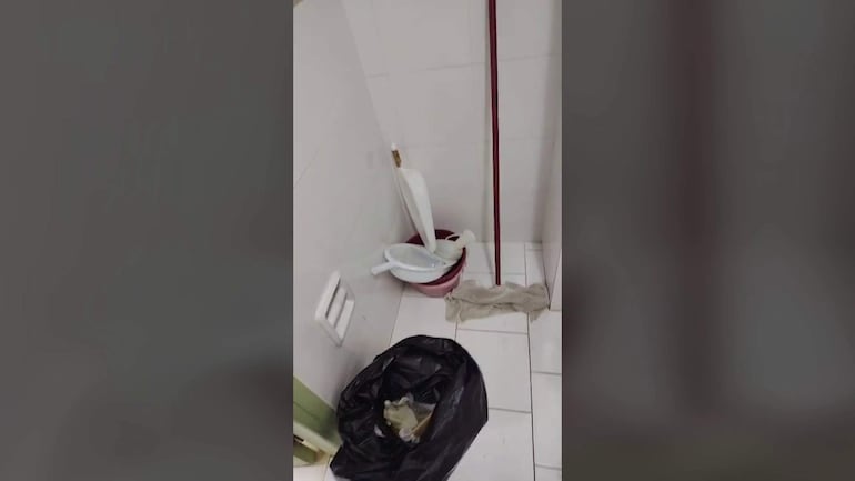 Baño del Hospital de Trauma con cubo de basura negro, balde rojo y toalla en el suelo, reflejando mala higiene.