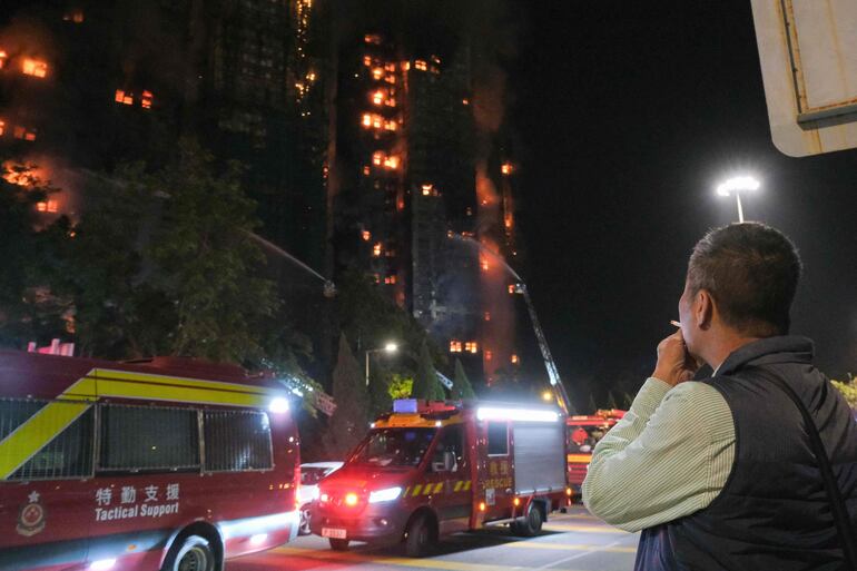 Un espectador fuma un cigarrillo cerca de los vehículos de rescate mientras un gran incendio arrasa varios bloques de apartamentos en la urbanización Wang Fuk Court (al fondo), en el distrito Tai Po de Hong Kong, el 26 de noviembre de 2025.