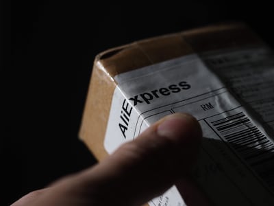Mano sosteniendo un paquete de AliExpress con fondo negro, en un ambiente cerrado y con iluminación tenue.