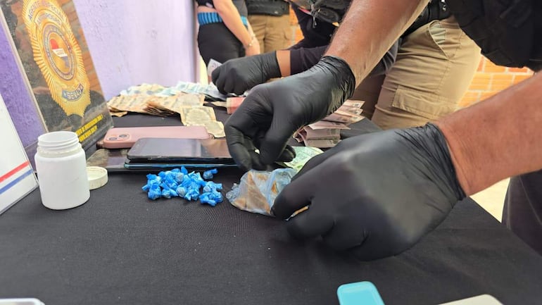 Manos con guantes manipulan objetos sobre tela negra, con billetes apilados y un frasco blanco en la mesa.