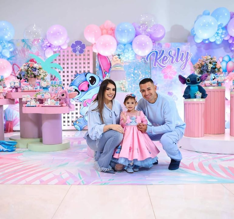 Niña en vestido rosa con peinado adornado, sonriendo mientras dos adultos la observan felizmente en un ambiente decorado con globos.