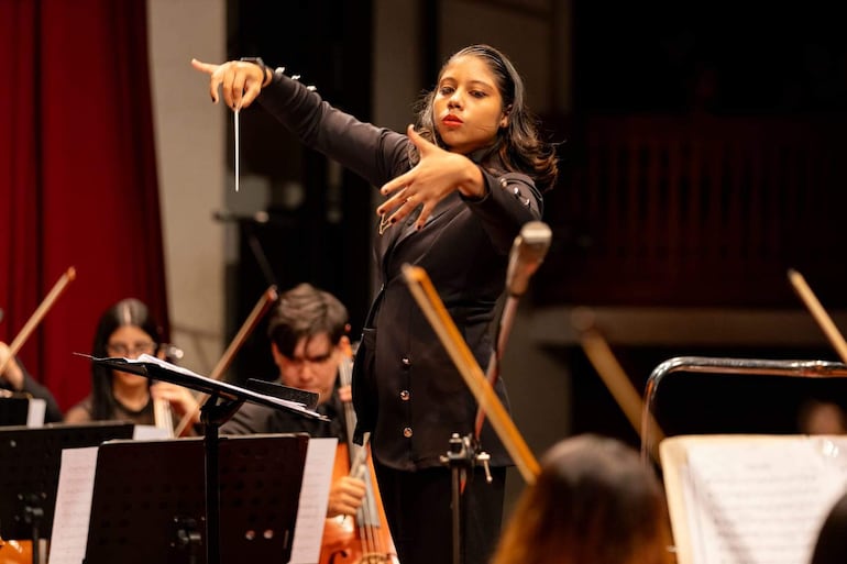 Directora de orquesta de pie en el escenario, vestida de negro, con expresión concentrada y batuta en mano, mientras músicos tocan detrás.