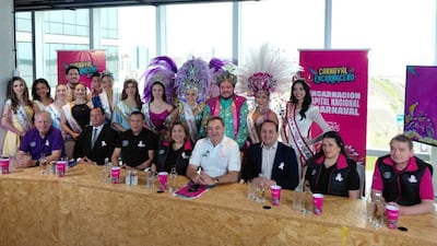 Mujeres con trajes de carnaval en el centro, mientras hombres en una mesa discuten el evento con entusiasmo.