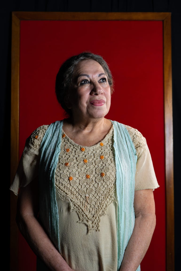 Mujer de cabello corto y canoso, vestida con blusa floral y chal azul, mirando contemplativa en un fondo rojo iluminado.