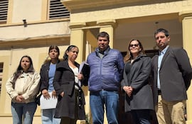 Grupo de seis personas, tres mujeres y tres hombres, de pie ante edificio oficial, algunos con documentos. Atmosfera seria y atenta.