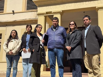 Grupo de seis personas, tres mujeres y tres hombres, de pie ante edificio oficial, algunos con documentos. Atmosfera seria y atenta.