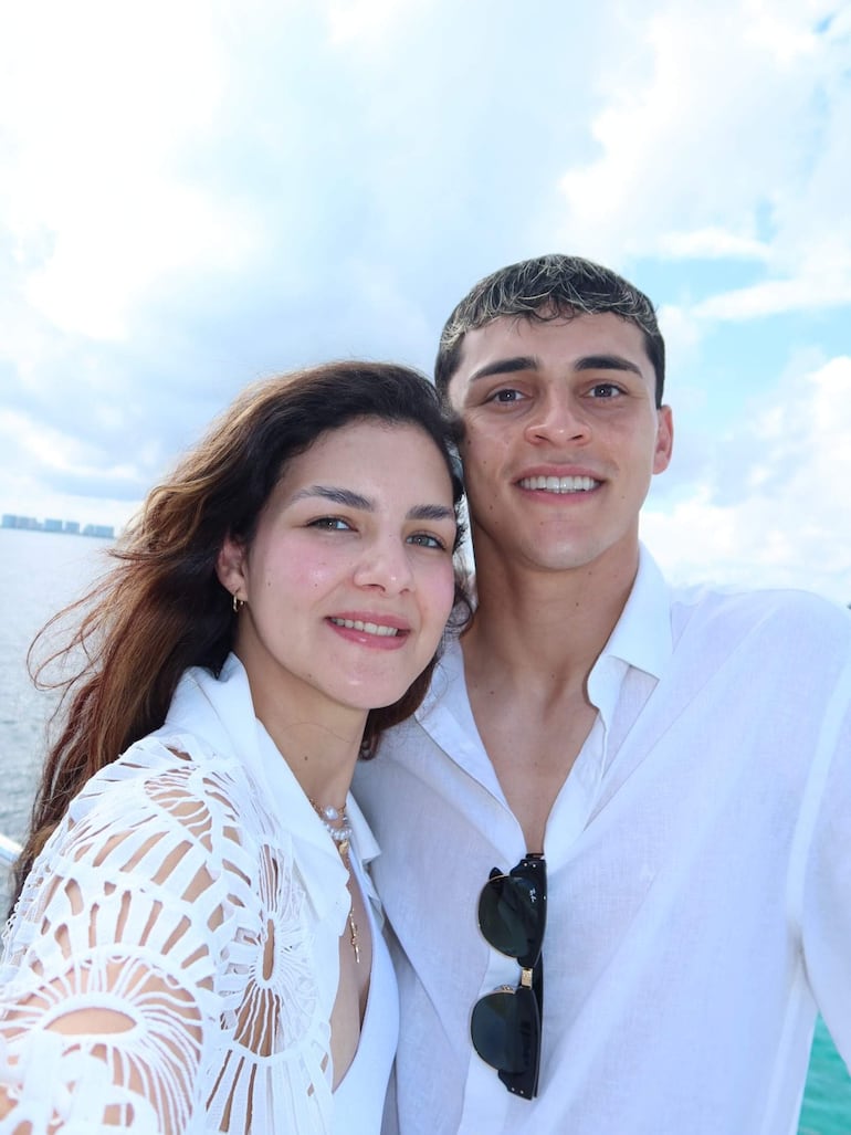 Hombre y mujer sonrientes posando para un selfie con agua de fondo y cielo nublado.