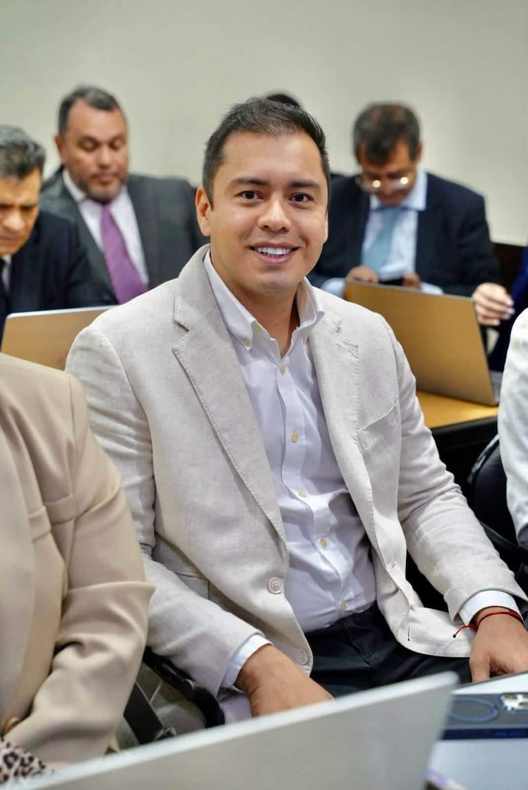 Miguel Prieto Vallejos sonríe, vestido con blazer claro y camisa blanca, rodeado de otros hombres en trajes oscuros en un ambiente profesional.