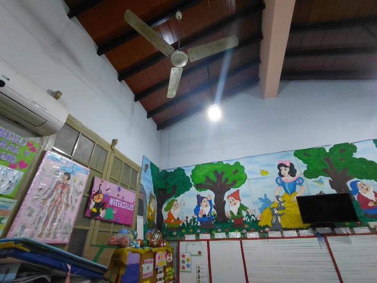 Aula con mural de Blancanieves, ventilador en el techo, aire acondicionado y estantes con materiales educativos.