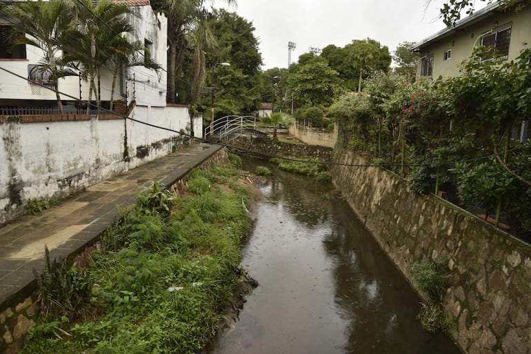 Canal de agua bordeado de vegetación y pavimento en un área urbana tranquila, con edificaciones y postes de luz al fondo.