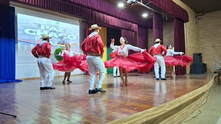 Cinco danzantes en trajes rojos bailan en el escenario, rodeados de un fondo decorado con 'Paz del Chaco'.