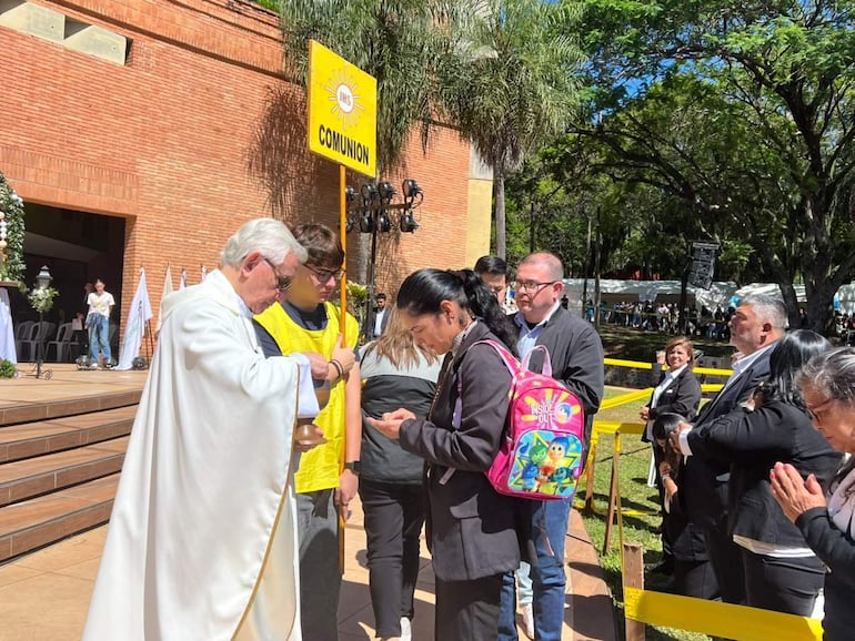 Sacerdote con vestiduras blancas realiza comunión, joven con camiseta amarilla sostiene cartel en ceremonia rodeada de asistentes en actitud de oración.
